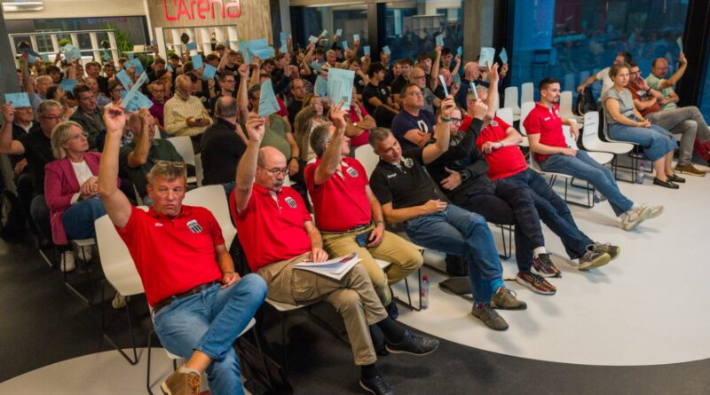 Viele Menschen sitzen in einem Raum und haben blaue Stimmkarten in der Hand. Die meisten von Ihnen heben sie gerade mit einer Hand in die Luft um für etwas abzustimmen.