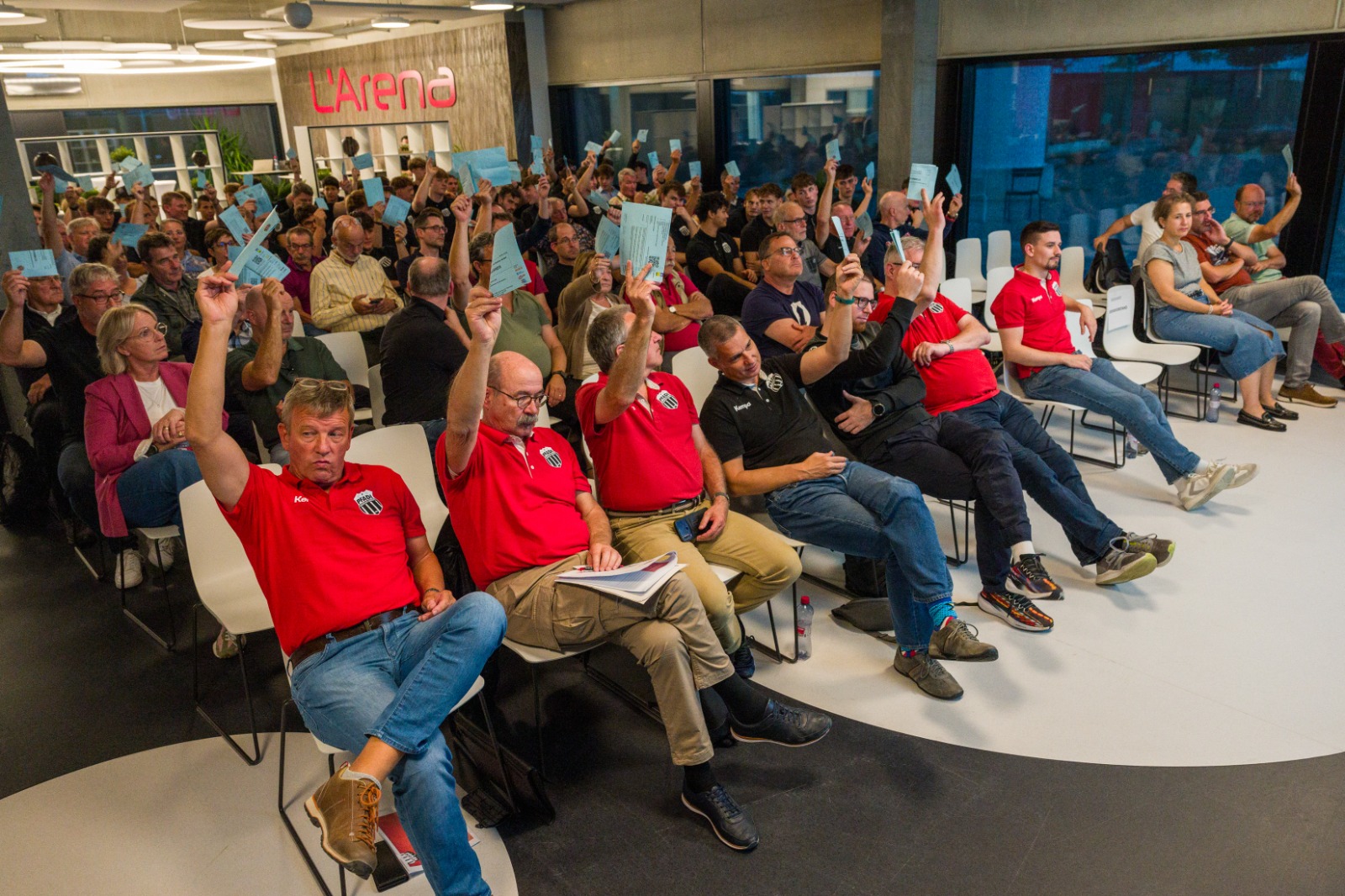 Viele Menschen sitzen in einem Raum und haben blaue Stimmkarten in der Hand. Die meisten von Ihnen heben sie gerade mit einer Hand in die Luft um für etwas abzustimmen.